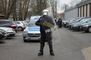 policjant ubrany w odzież ochronną i z tarczą w tle radiowóz policyjny