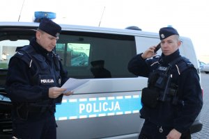 policjanci na tle radiowozu. jeden z nich trzyma w dłoni białą kartkę drugi trzyma przy uchu telefon komórkowy