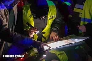 fotografia przedstawia torsy policjantów pochylonych nad mapą. Jeden z mężczyzn trzyma w dłoni latarkę i oświetla mapę, która leży na masce samochodu. Panuje mrok .